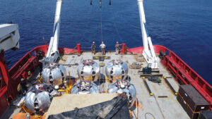 Le unità di rivelazione (stringhe) di KM3NeT-ARCA a bordo della nave Optimus Prime, pronte per essere deposte a 3500 m di profondità. Le stringhe sono avvolte su un lanciatore sferico galleggiante e connesse a una base metallica (pitturata in giallo) che funge da ancora. Il verricello della nave le posa delicatamente sul fondo sotto la supervisione di un robot, che le connette alla rete sottomarina e attua il meccanismo di sgancio del lanciatore.